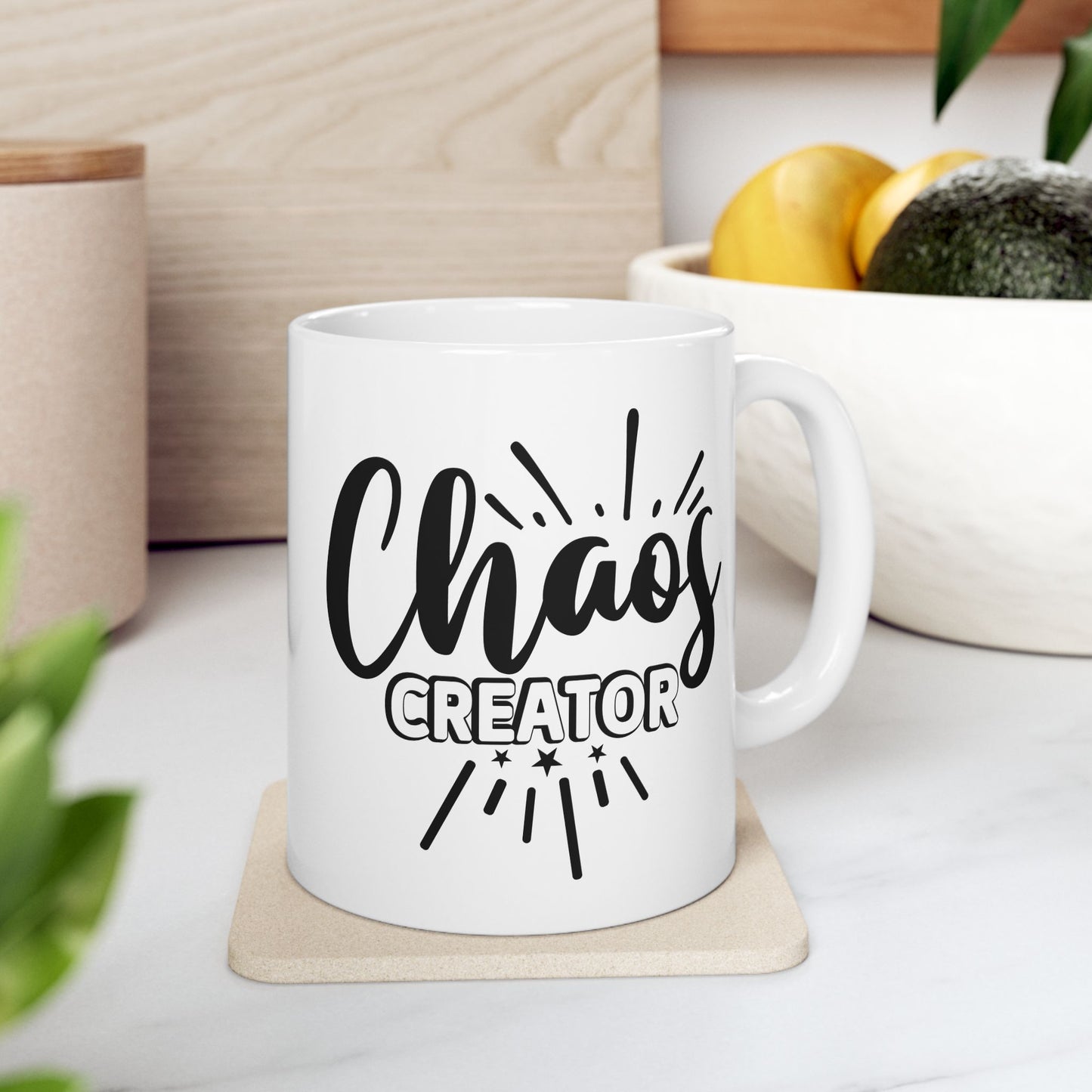 White mug with 'Chaos Creator' text on a wooden coaster, surrounded by plants and a bowl of fruit.
